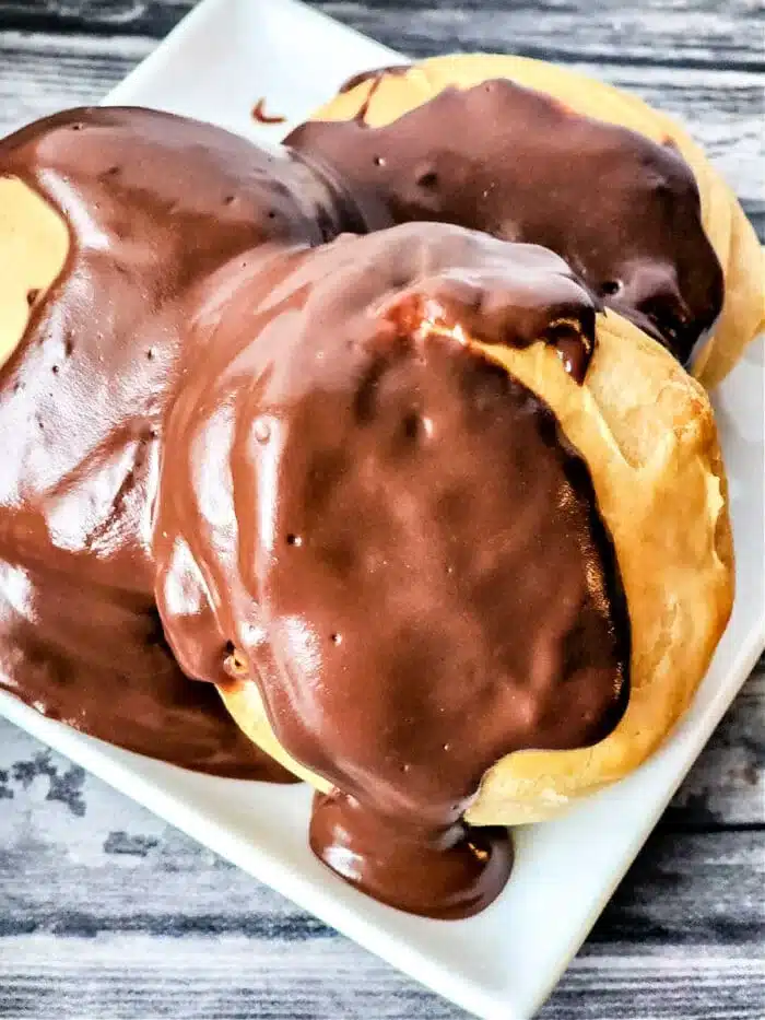 Three cream puffs covered in thick chocolate glaze are displayed on a white rectangular plate, showcasing the richness akin to mastering how to make chocolate gravy. The background consists of a wooden surface with a gray and black grain pattern.