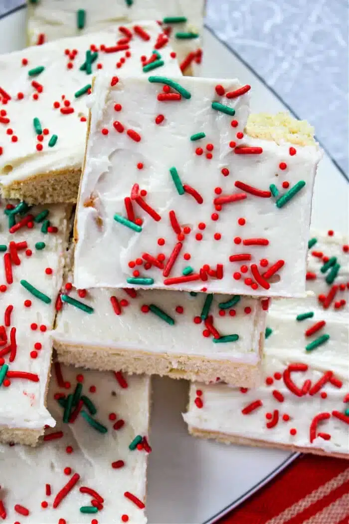 A white plate holds a stack of frosted Christmas bar cookies. The cookies are topped with white frosting and decorated with red and green sprinkles. There is a red and white striped cloth partially visible at the bottom.