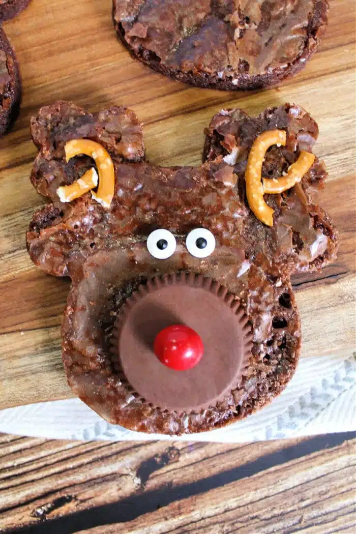 A reindeer-shaped Christmas brownie with a peanut butter cup for a nose, a red candy on top, candy eyes, and broken pretzels as antlers rests on a wooden surface. Other similar brownies are partially visible in the background.