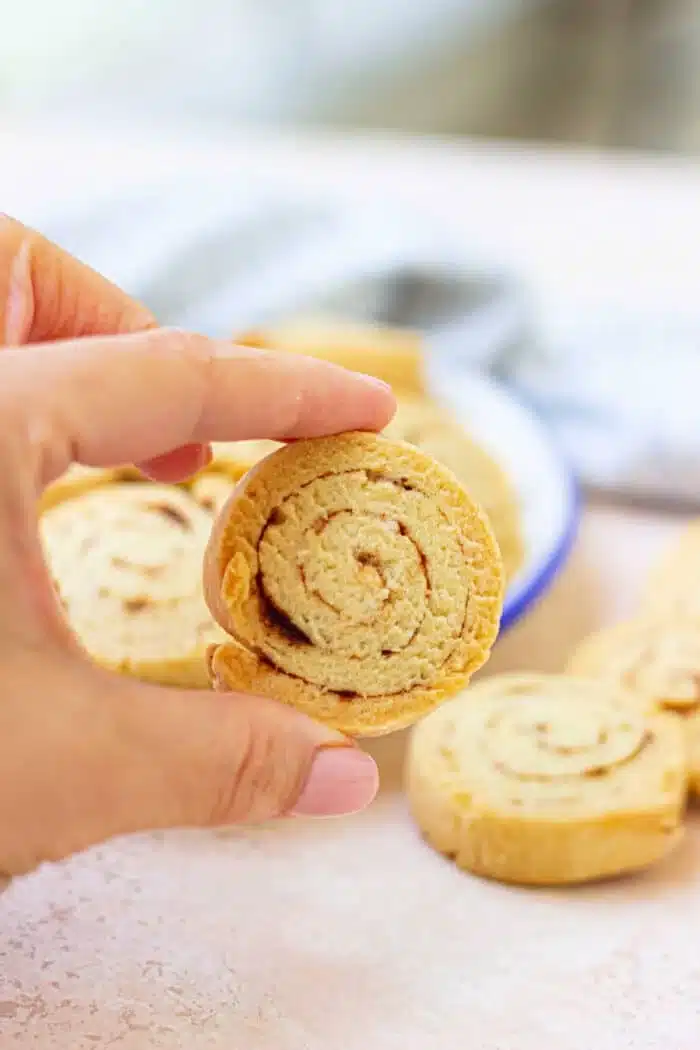 A hand holding a round, swirled slice of homemade shortbread cookie. More cinnamon biscuits of the same type are visible in the background on a white surface, with a blue-rimmed dish partially visible holding some cookies.