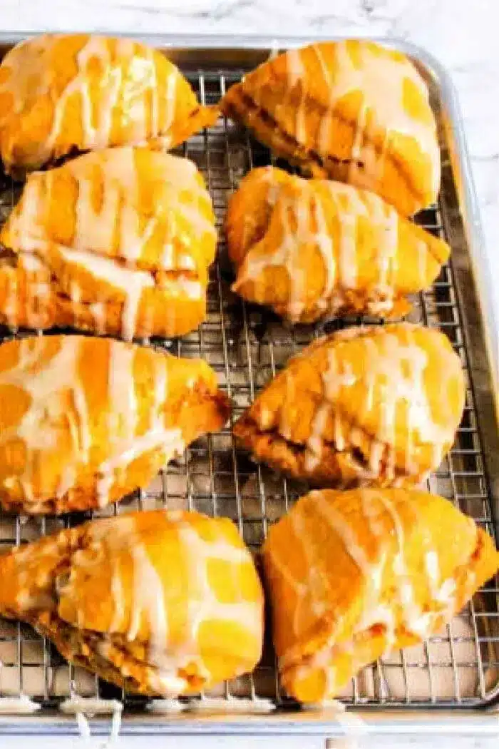 Eight golden brown pastries, reminiscent of a Starbucks pumpkin scone, with glaze drizzle are arranged on a cooling rack placed over a baking sheet.