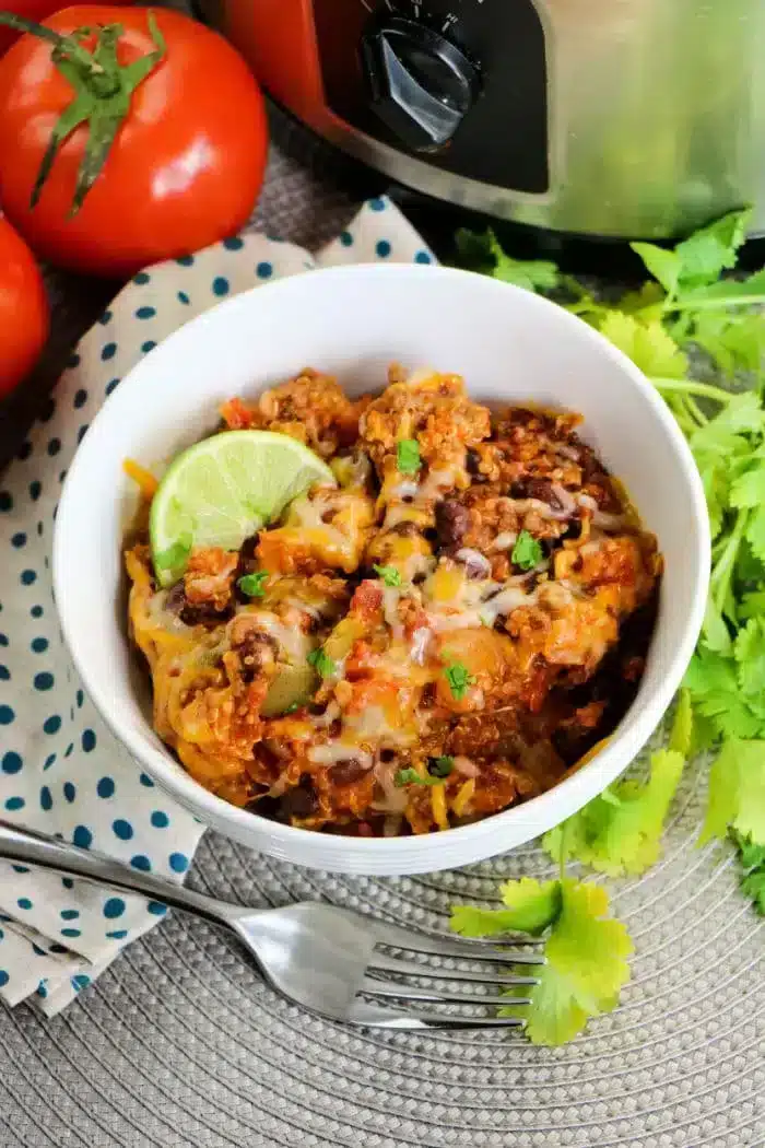 A white bowl filled with chili topped with melted cheese, chopped cilantro, and a lime wedge. Beside the bowl are a fork, fresh cilantro leaves, whole tomatoes on the vine, a polka-dotted cloth, and a slow cooker in the background hinting at a crockpot enchilada casserole in progress.