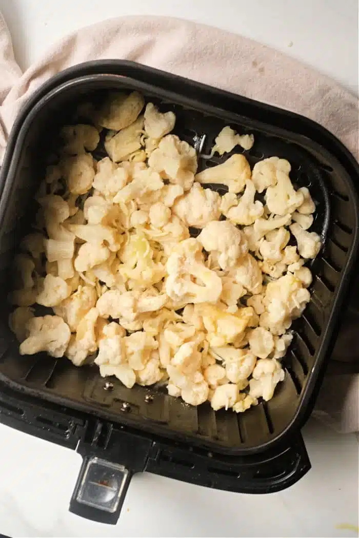 Frozen cauliflower florets in an air fryer basket rest delicately on a light-colored surface, ready to crisp to perfection.