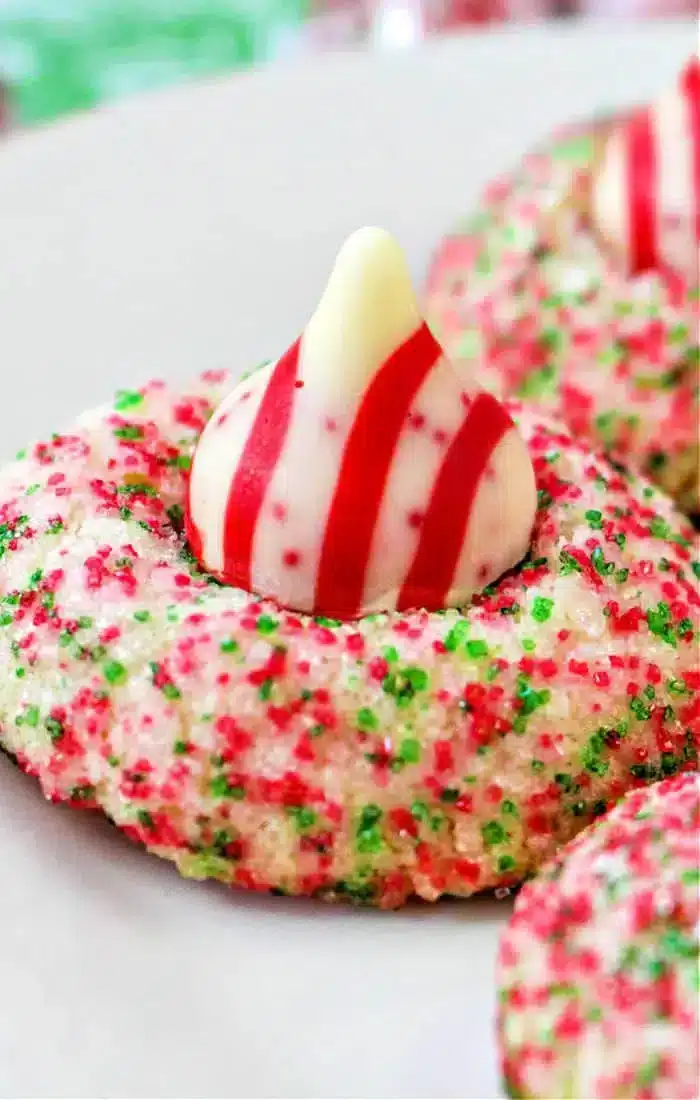 A festive cookie covered in red and green sugar sprinkles features a striped red and white Hershey Kiss shaped like a drop on top. Perfect for Christmas, this treat sits elegantly on a white plate.