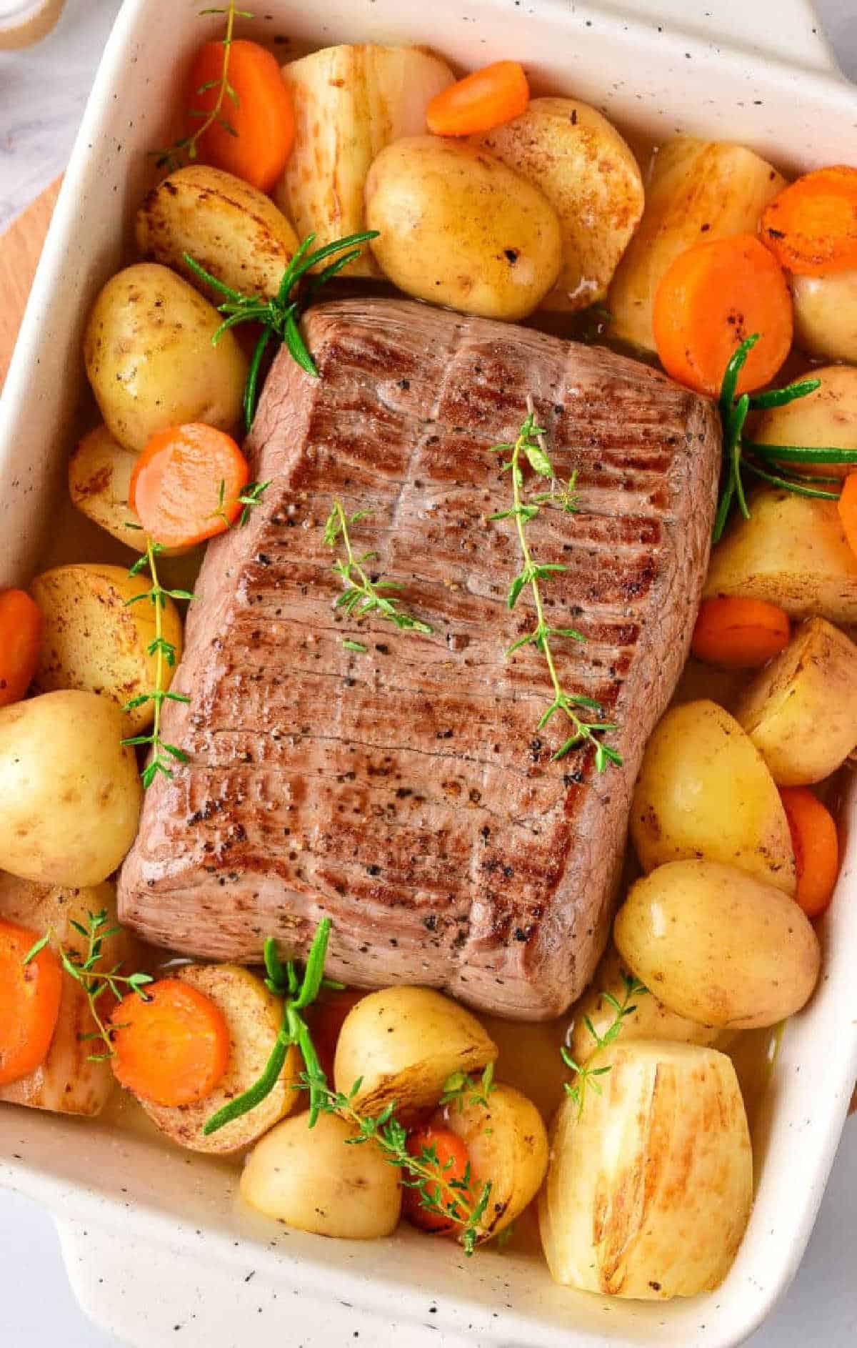 Roast beef with potatoes, carrots, and herbs in a white baking dish—perfect for learning how long to cook chuck roast in oven at 350 for a tender, flavorful meal.