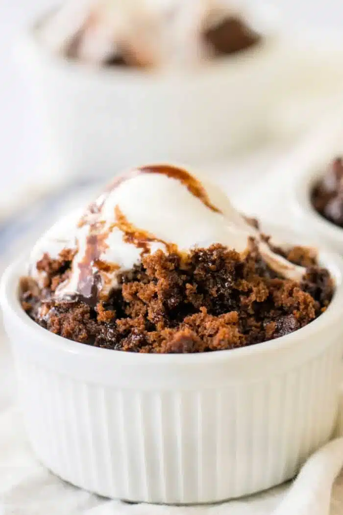 A white ramekin filled with a delectable chocolate brownie cake topped with a scoop of vanilla ice cream and drizzled with rich chocolate syrup. Another ramekin with a similar treat is visible in the blurred background.