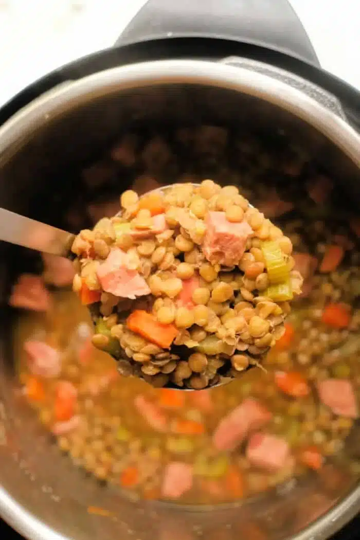 A ladle holds a serving of lentil soup with ham, showcasing visible chunks of meat, carrots, and celery, above a pot brimming with more soup. The contents display a delightful mix of earthy and colorful ingredients.