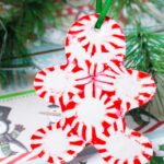 A Christmas ornament shaped like a gingerbread man is made from red and white peppermint candies, creating a whimsical, melting peppermint candy effect. It hangs by a green ribbon, with green pine branches in the background and holly designs on paper beneath it.