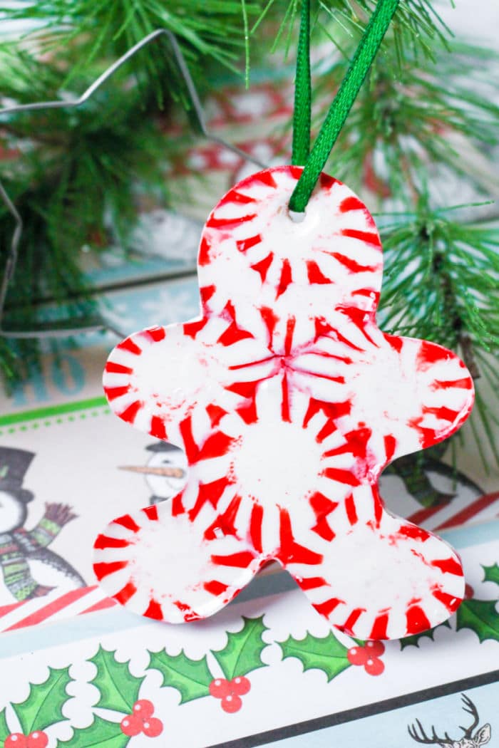 A Christmas ornament shaped like a gingerbread man is made from red and white peppermint candies, creating a whimsical, melting peppermint candy effect. It hangs by a green ribbon, with green pine branches in the background and holly designs on paper beneath it.