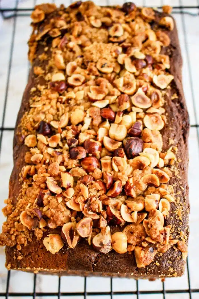 A loaf cake topped with a crumbly mix of chopped nuts, including hazelnuts, reminiscent of Nutella bread, sits on a cooling rack. The golden-brown cake has a textured top with scattered pieces of nuts and crumbs visible.