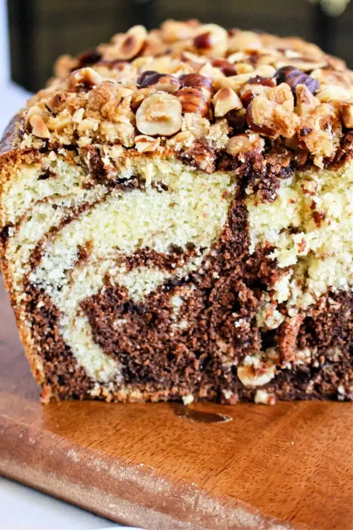 A close-up of a sliced marble Nutella loaf cake on a wooden surface. The cake features a mix of chocolate and vanilla swirls, topped with chopped nuts and a streusel-like crumble.