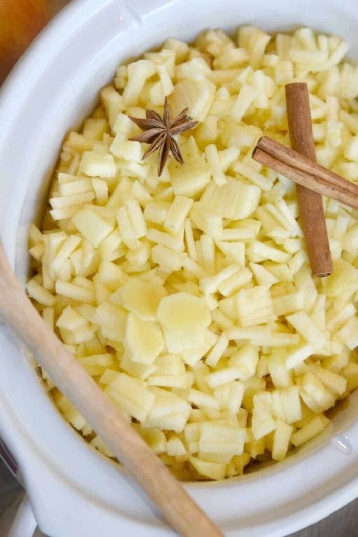 A white bowl filled with diced apples, ready to transform into slow cooker apple butter. A star anise and a cinnamon stick are placed on top of the apples, while a wooden spoon rests on the edge of the bowl.