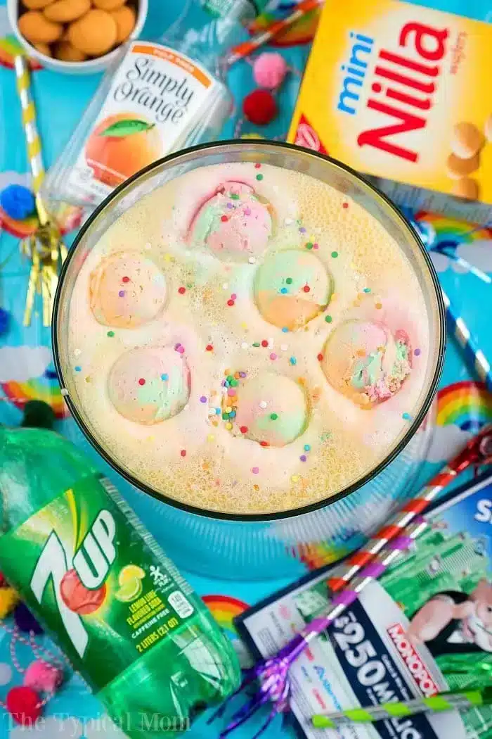 A bowl of colorful party punch with scoops of rainbow sherbet and sprinkles is surrounded by cans of Simply Orange and 7Up, a box of Mini Nilla wafers, and packets of Kool-Aid. Straws and festive napkins add the perfect touch to this delightful setup.