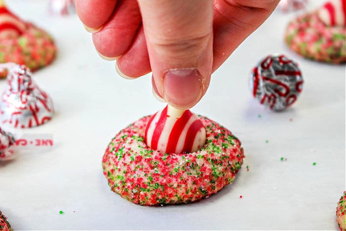 A hand is pressing a peppermint-striped candy into the center of a round, sugar-coated cookie. The cookie, reminiscent of Peanut Butter and Grape Jelly Cookies in its creativity, is covered in red and green sugar sprinkles with more decorated cookies and candies visible in the background.