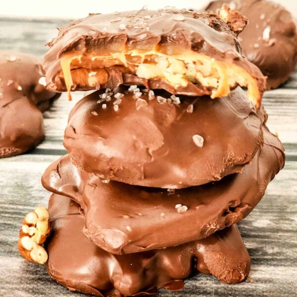 A stack of chocolate-covered caramel and pecan turtles is shown. The top cluster has a bite taken out of it, revealing luscious caramel and crunchy pecans. They are sprinkled with sea salt and sit invitingly on a wooden surface.