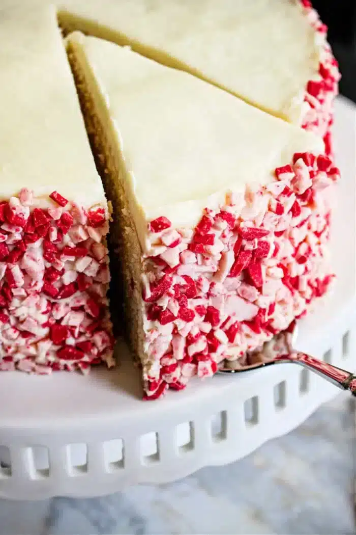 A Peppermint Cake with white frosting and red and white crushed candy on the sides is displayed on a white cake stand. A slice has been cut and is being removed with a cake server.