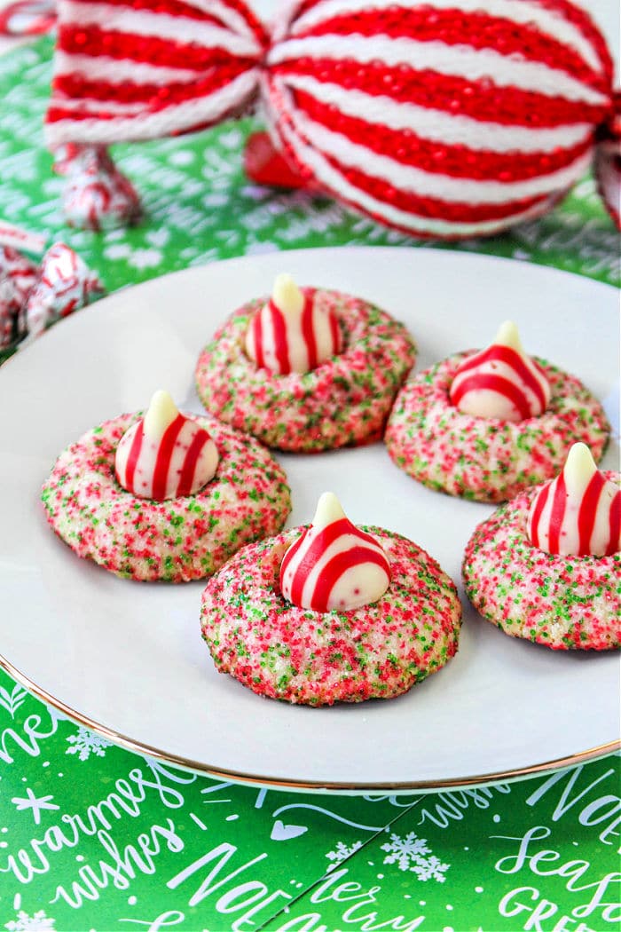 A plate with five holiday-themed cookies topped with red and white striped candies, reminiscent of Hershey Kiss Christmas Cookies. The cookies are coated in vibrant red and green sprinkles. In the background, a large, wrapped decorative item boasts festive red and white stripes on a jolly green surface.