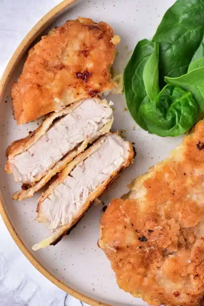 A plate featuring breaded and fried pork cube steaks, with one chop sliced to show the tender interior. Fresh spinach leaves are placed beside the chops on the light-colored plate.