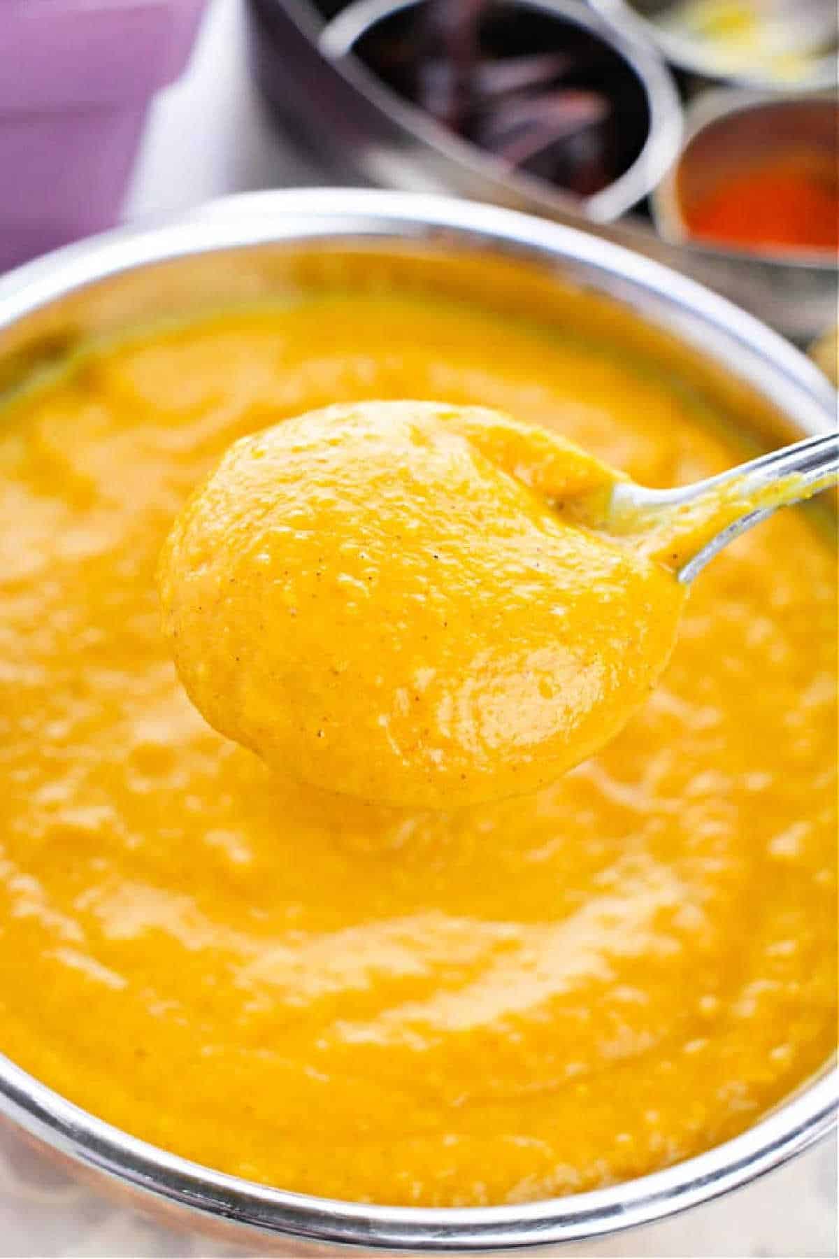 A spoon holding thick, creamy Pumpkin Soup with Canned Pumpkin hovers over a bowl filled with the same golden curry sauce.