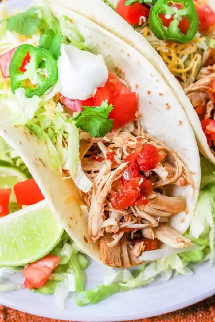 Close-up of two Rotel Tacos filled with shredded meat, diced tomatoes, lettuce, cheese, and jalapeño slices, garnished with cilantro and a dollop of sour cream. Lime wedges are placed beside the tacos.