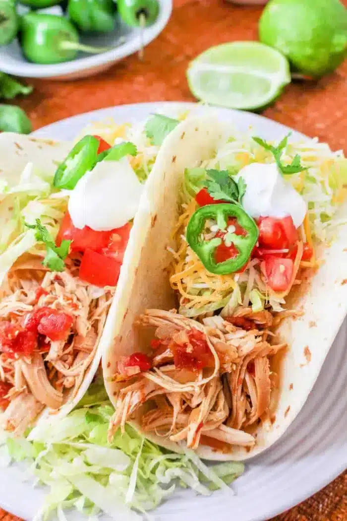 Two Rotel tacos filled with shredded chicken, diced tomatoes, shredded cheese, jalapeño slices, sour cream, and lettuce on a white plate. A halved lime and extra jalapeños are in the background.