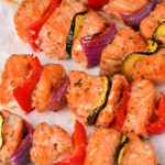 Close-up of salmon kabobs on a white surface. The skewers include chunks of salmon, red bell peppers, red onions, and zucchini, all lightly seasoned. Garnished with sprigs of fresh parsley.