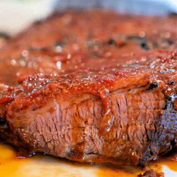 Close-up of a Crockpot Brisket Recipe with a glistening, caramelized exterior. The meat appears juicy and tender, with a portion cut to show its texture. A colorful, blurred background contrasts with the rich, reddish-brown tones of the brisket.