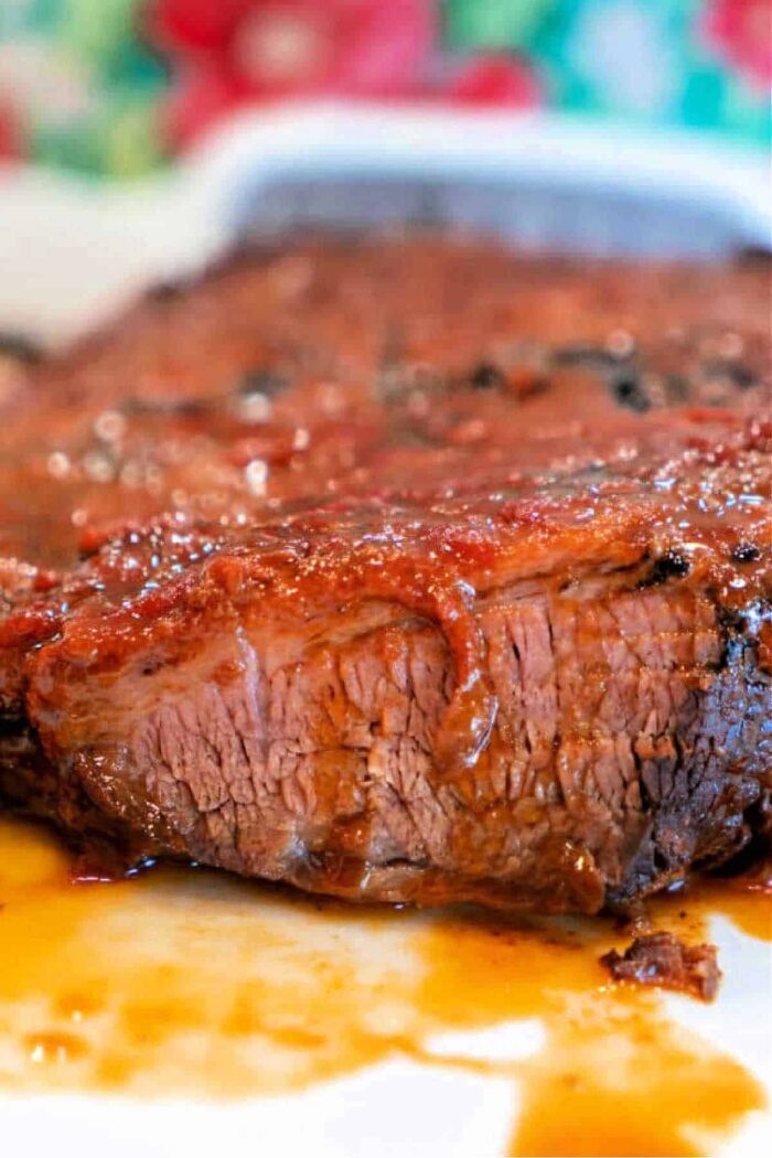 Close-up of a Crockpot Brisket Recipe with a glistening, caramelized exterior. The meat appears juicy and tender, with a portion cut to show its texture. A colorful, blurred background contrasts with the rich, reddish-brown tones of the brisket.