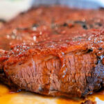 Close-up of a sliced piece of cooked brisket from a Crockpot Brisket Recipe. The meat has a slightly charred, caramelized exterior with a reddish-brown sauce coating, and is sitting in a small pool of juices on a white plate. A colorful, out-of-focus background is visible.