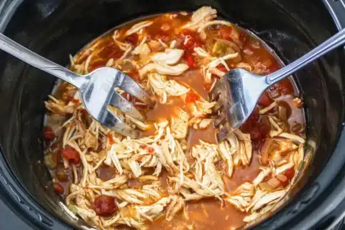 Two forks are shredding cooked chicken in a slow cooker. The chicken mingles with a vibrant Rotel sauce featuring diced tomatoes and green peppers, perfect for mouth-watering tacos.