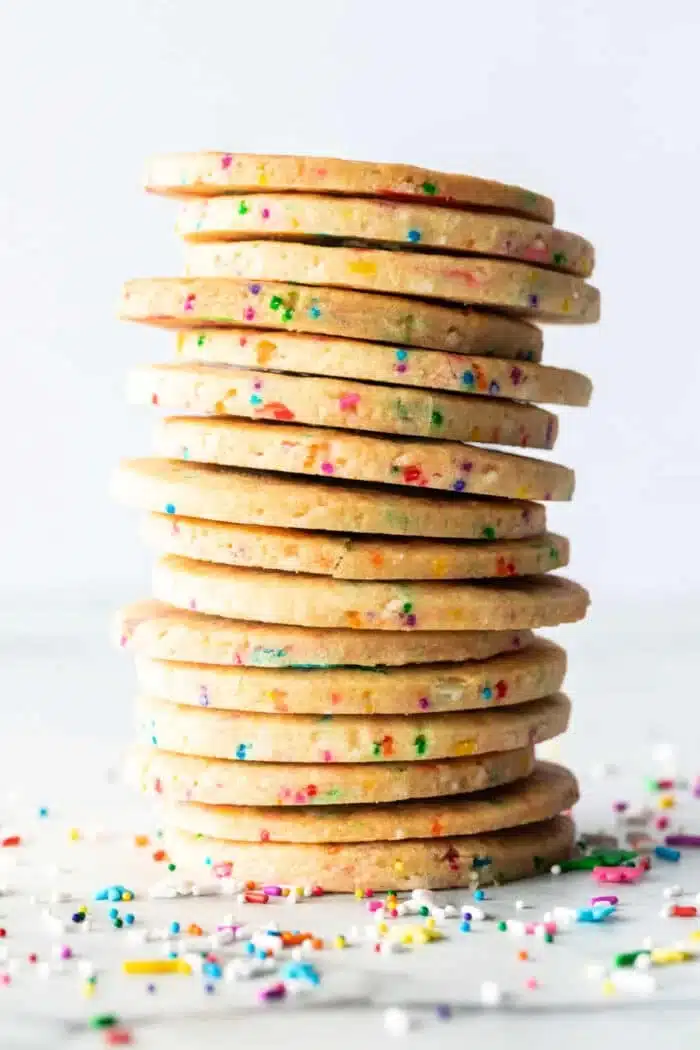 A stack of round Sprinkle Sugar Cookies with colorful sprinkles embedded throughout is displayed against a plain white background. Additional sprinkles are scattered at the base of the stack.