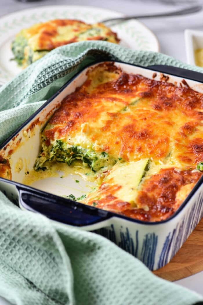 Baked spinach and cheese casserole in a ceramic dish with a piece removed, reminiscent of vegetable lasagna with white sauce, topped with golden melted cheese.
