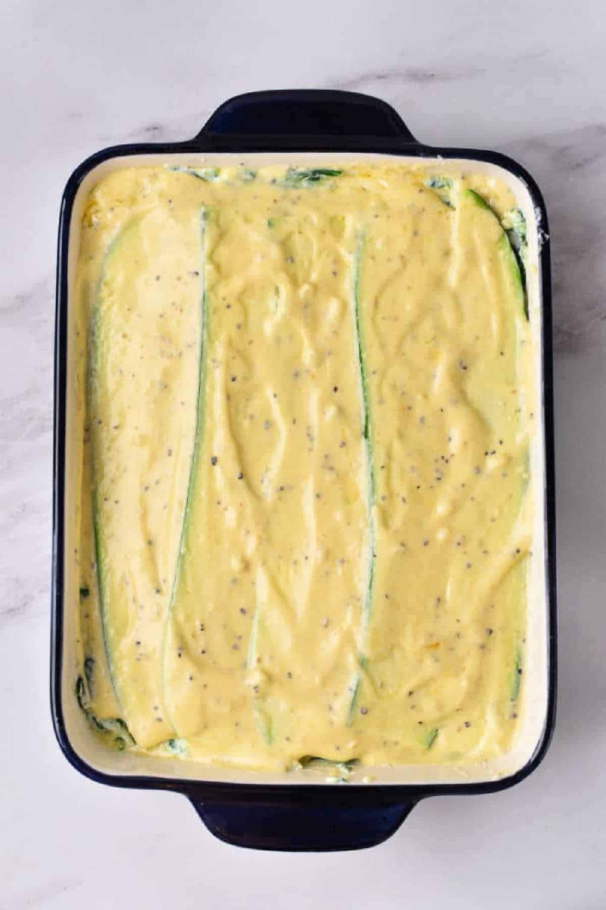 A baking dish filled with an unbaked, creamy, yellow mixture and layered strips of zucchini, ready to become a delicious vegetable lasagna with white sauce.