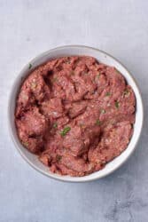 Ground venison mixed with herbs in a white bowl on a gray surface.