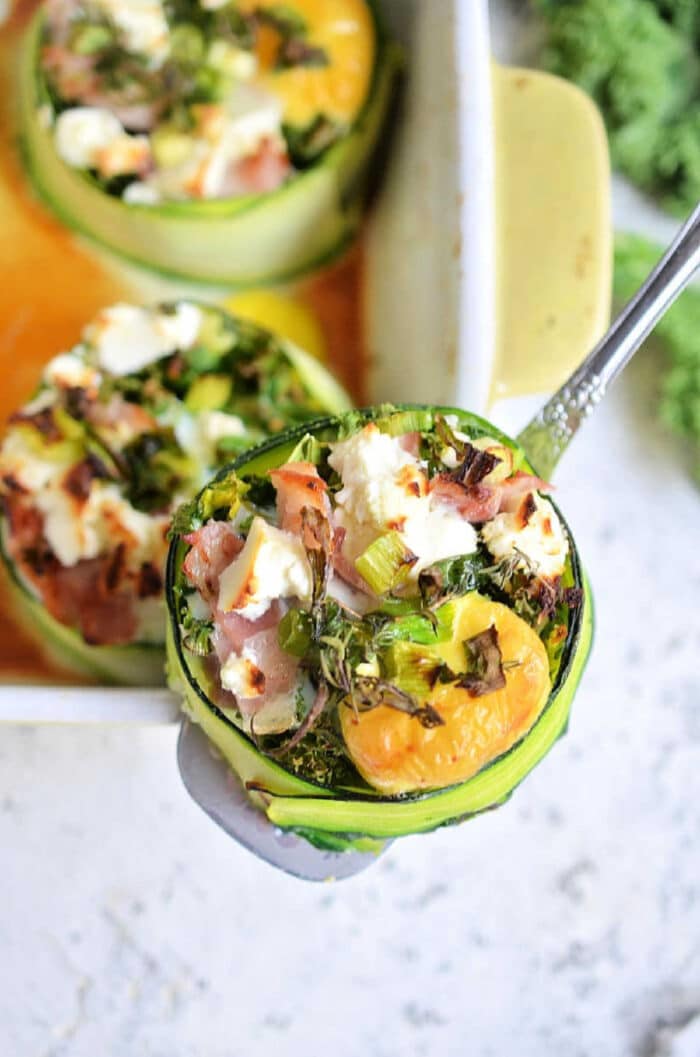 A close-up of a dish showcasing stuffed zucchini cups filled with eggs, greens, ham, and cheese. One zucchini cup is being lifted with a silver spatula, revealing a colorful and neatly arranged filling. A baking dish with more eggs in the oven-stuffed zucchini cups is partially visible.