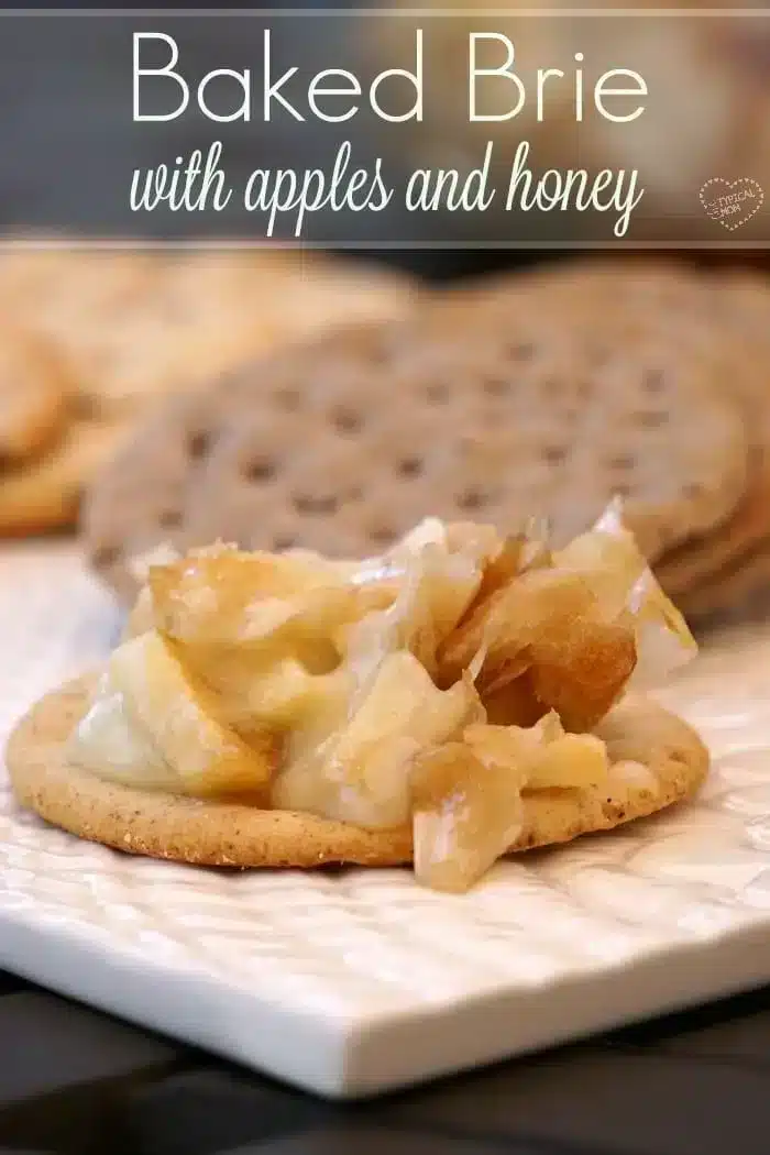 A cracker topped with easy baked Brie cheese, apple pieces, and honey sits on a white plate. Blurred crackers appear in the background. The text "Baked Brie with apples and honey" graces the top.