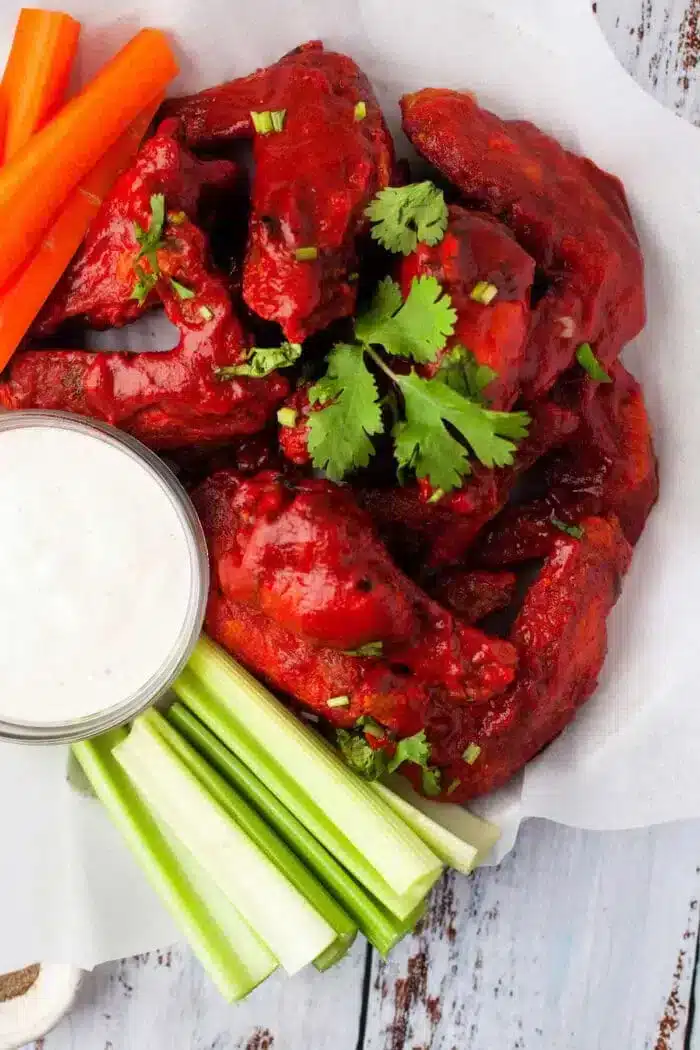 A plate of saucy buffalo chicken wings, cooked to perfection in an air fryer, garnished with cilantro, served with carrot and celery sticks, and a small cup of white dipping sauce on the side. The food is presented on parchment paper.