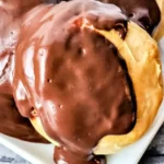 Close-up of two biscuits topped with a luscious layer of chocolate glaze reminiscent of chocolate gravy on biscuits, placed on a white plate. The background features a wooden surface.