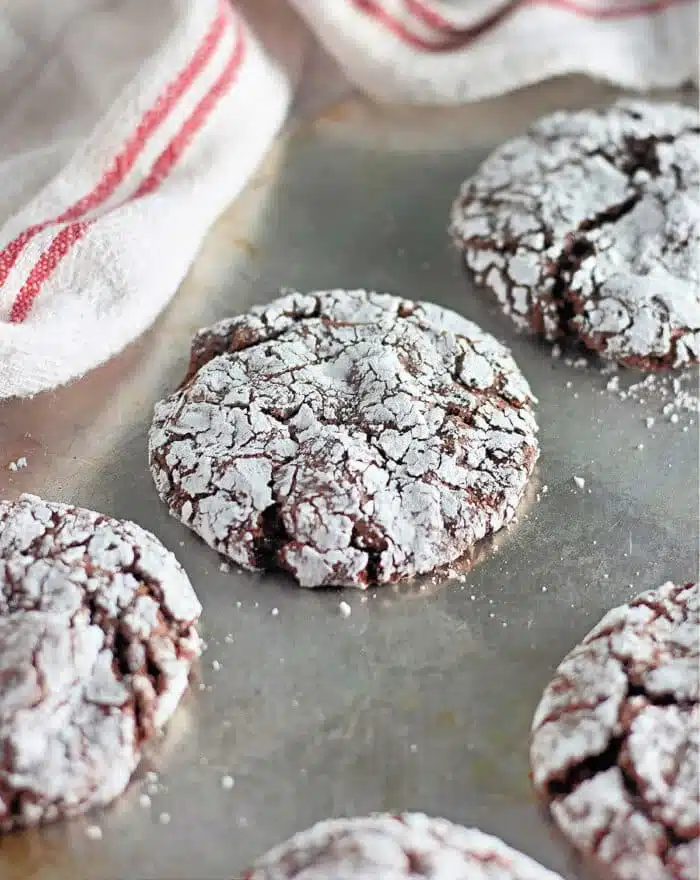 A close-up of chocolate crinkle cookies on a baking sheet. The cookies are cracked on top and dusted with powdered sugar, revealing the chocolate underneath. Freshly baked as toaster oven cookies, a white towel with red stripes is partially visible in the background.