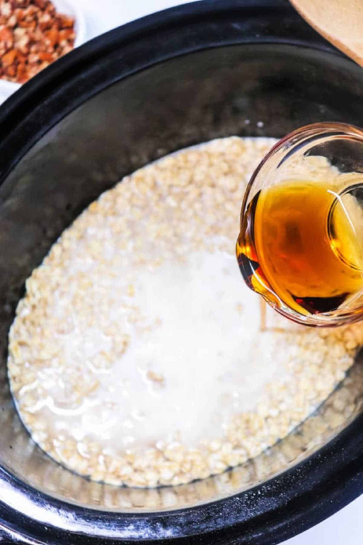 A hand pours a golden liquid into a slow cooker filled with oats and milk, preparing delicious Cinnamon Oatmeal.
