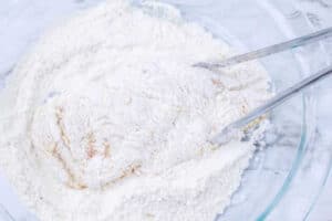 Raw chicken breast being coated in flour with metal tongs in a glass bowl on a marble surface, ready to become an Air Fryer McDonalds Fried Chicken Sandwich.