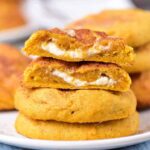 A close-up shows a stack of Pumpkin Cream Cheese Cookies with a luscious cream cheese filling. The top cookie is cut in half, revealing the rich, creamy layer inside. The cookies are arranged on a white plate, placed on a blue tablecloth.