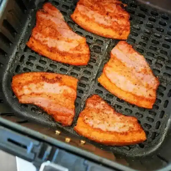 Five crispy, browned pieces of pork belly bacon are inside an air fryer basket. The bacon pieces are arranged in a single layer, showing the ridged marks from the basket's surface. The air fryer is open, revealing the cooked pork belly bacon inside.