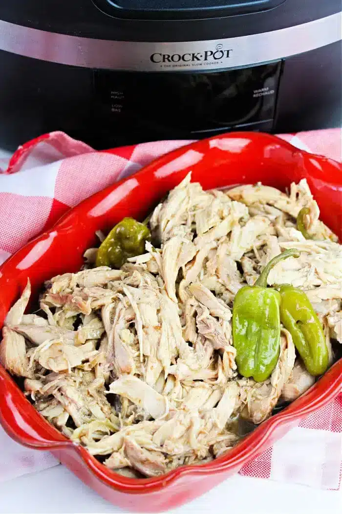 A red dish filled with shredded pepperoncini chicken and whole green peppers is placed on a white and red checkered cloth. In the background, there is a black Crock-Pot slow cooker.