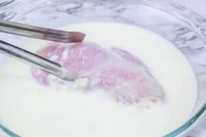 Raw chicken breast being dipped in a bowl of milk with metal tongs on a marble surface, the first step to recreating an Air Fryer McDonalds Fried Chicken Sandwich at home.