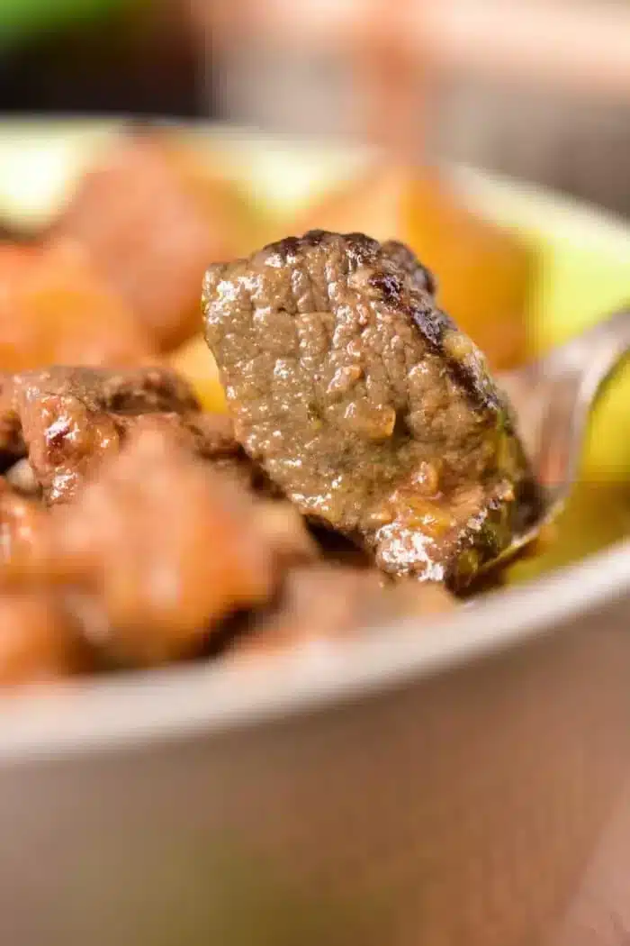 A close-up image of a spoon lifting a piece of cooked venison from a dish. The meat is part of a hearty venison stew, with other pieces visible in the blurred background. The meat is brown and looks well-cooked, inviting you to try this delicious venison stew recipe.