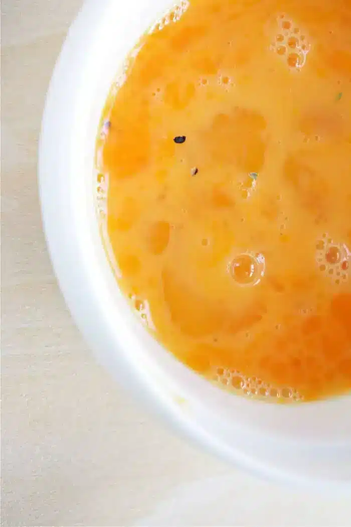 A close-up view of a bowl filled with an orange liquid, likely part of an egg wash recipe. The liquid appears smooth with a few small bubbles and specks on its surface. The bowl is white and sits on a light wooden surface.