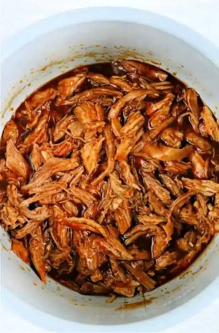 A pot filled with shredded pulled pork butt, perfectly cooked in an Instant Pot. The meat is thoroughly mixed with a rich, reddish-brown sauce, giving it a glossy appearance. Each strand of pork is evenly coated in the luscious sauce.