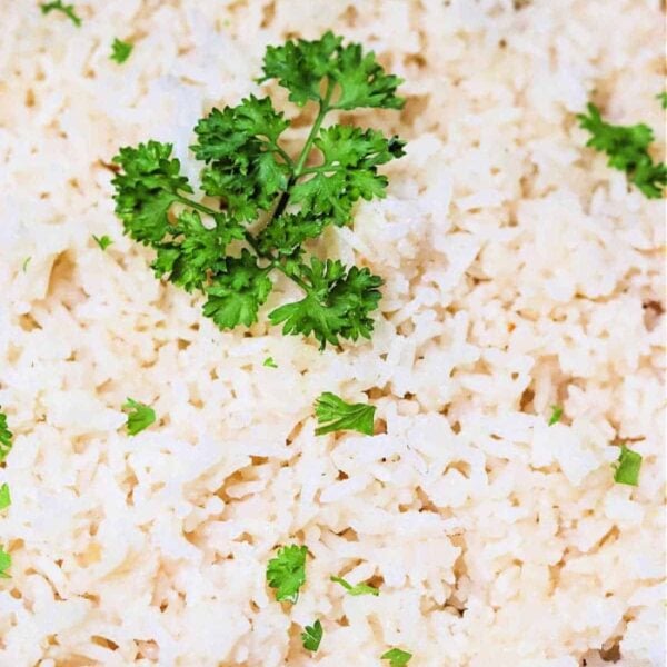 A close-up image of a dish of cooked white rice, prepared in a slow cooker, garnished with a sprig of fresh parsley and small parsley pieces scattered throughout.