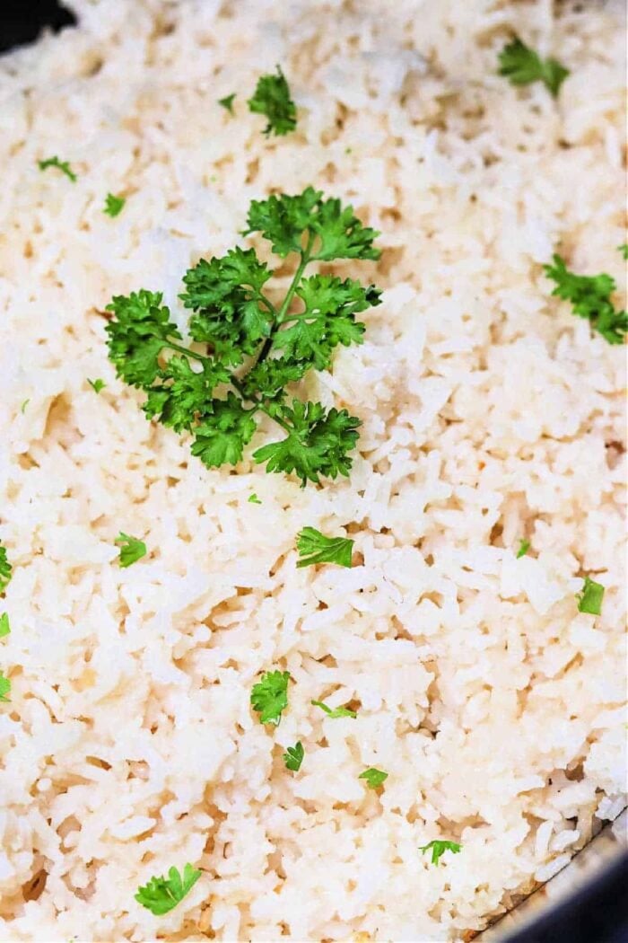 A close-up image of a dish of cooked white rice, prepared in a slow cooker, garnished with a sprig of fresh parsley and small parsley pieces scattered throughout.